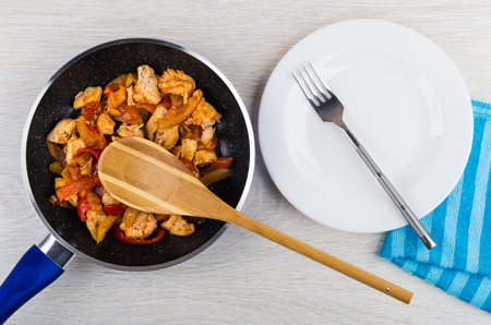 Bamboo spoon in frying pan with fried chicken meat and sweet pepper, empty plate, fork on napkin on wooden table. Top viewの写真素材