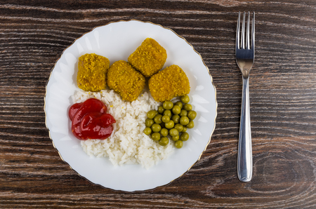 Nuggets from turkey with rice, green peas, ketchup in plate, fork on wooden table. Top viewの写真素材