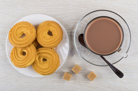 Cocoa with milk in cup, spoon on saucer, cookies in saucer, sugar cubes on wooden table.Top viewの写真素材