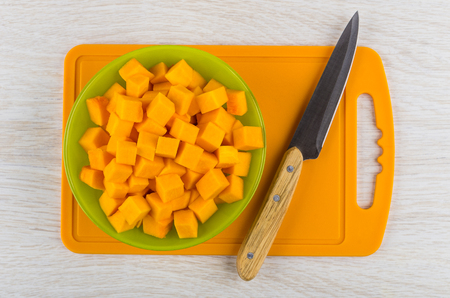 Cubes of raw pumpkin in green bowl, kitchen knife on cutting board on wooden table. Top viewの写真素材