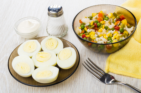 Halves of boiled eggs in plate, fork, hawaiian vegetable mix in brown bowl, napkin, mayonnaise, salt on wooden tableの写真素材