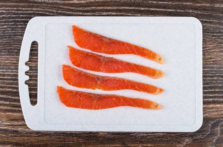 Slices of salted salmon on plastic cutting board on dark wooden table. Top view