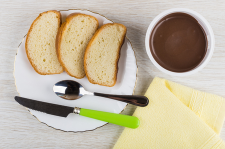 Jar of chocolate paste, pieces of bread, spoon, knife in plate, napkin on wooden table. Top viewの写真素材