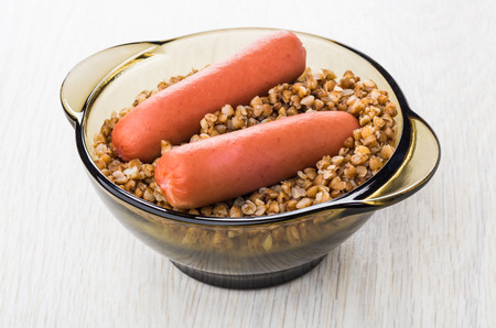Brown transparent bowl with boiled buckwheat and sausages on wooden tableの写真素材