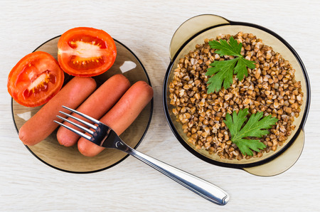 Brown transparent bowl with buckwheat, saucer with sausages an tomatoes, fork on wooden table. Top viewの写真素材