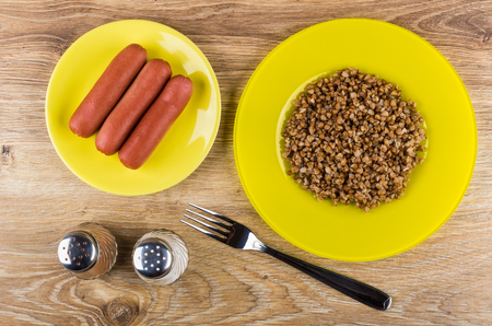 Boiled sausages in saucer, yellow plate with boiled buckwheat, salt, pepper, fork on wooden table. Top viewの写真素材