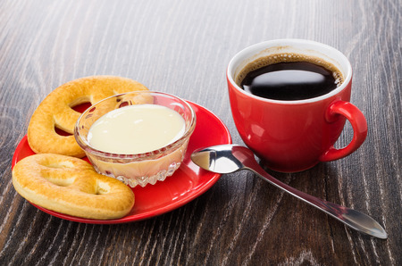 Cookies in form pretzels, condensed milk in bowl on saucer, spoon, cup of coffee on wooden tableの写真素材