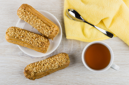 Eclairs in saucer, teaspoon on yellow napkin, cup of tea on wooden table. Top view