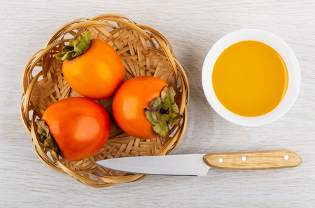 Three raw persimmon, kitchen knife in wicker basket, bowl with honey on wooden table. Top viewの写真素材