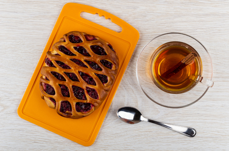 Pie with cherry filling on cutting board, cup of tea with cinnamon on saucer, teaspoon on wooden table. Top viewの写真素材