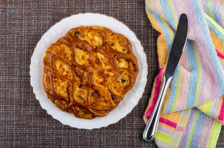Savory pie with filling in white plate, table knife on napkin on dark mat. Top view