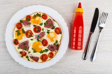 Fried eggs with sausages and tomato in white dish, bottle of ketchup, knife, fork on wooden table. Top viewの写真素材