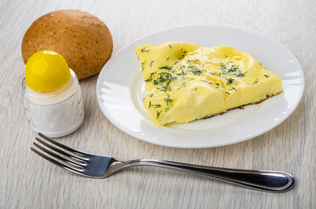 Piece of omelette with dill in plate, salt shaker, round bun, fork on wooden tableの写真素材
