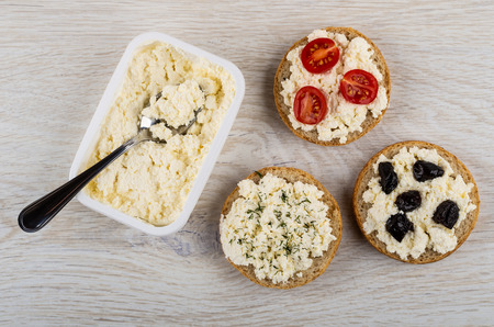 Ricotta cheese, spoon in white plastic box, sandwiches with cheese, dill, tomato, prunes on wooden table. Top viewの写真素材