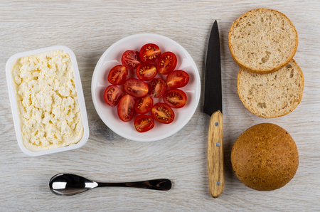 Ricotta cheese in white box, pieces of bread, bun, spoon, saucer with tomatoes, kitchen knife on wooden table. Top viewの写真素材