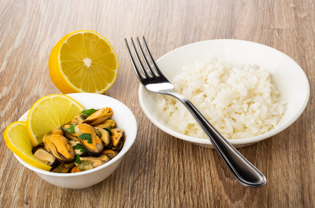 Boiled rice, fork in plate, half of lemon, cooked mussels with parsley in bowl on wooden tableの写真素材
