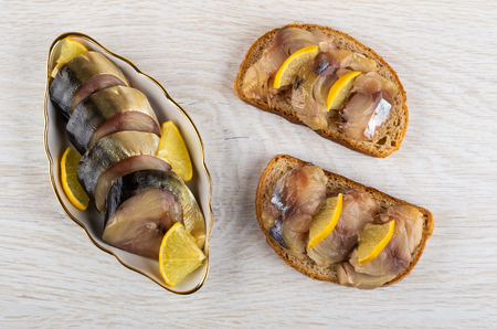 Pieces of smoked mackerel with slices of lemon in bowl, sandwiches with fish on wooden table. Top viewの写真素材