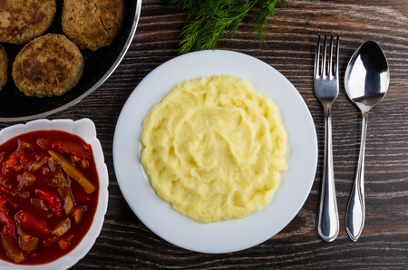 Fried cutlets in frying pan, bowl with lecho, dill, mashed potato in plate, fork, spoon on wooden table. Top viewの写真素材