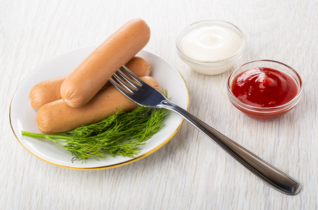 Boiled sausages, dill in white plate, sausage on fork, bowls with mayonnaise and ketchup on wooden tableの写真素材