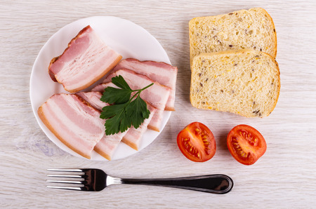 Piece and slices of brisket, parsley in white plate, pieces of bread, tomatoes, fork on wooden table. Top viewの写真素材
