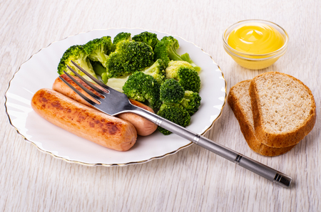 Boiled broccoli with fried sausages in plate, fork, mayonnaise in bowl, pieces of bread on wooden tableの写真素材