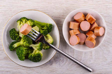 Boiled broccoli in plate, bowl with pieces sausages, fork on wooden table. Top viewの写真素材
