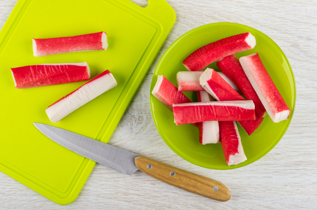 Crab sticks in green glass bowl, crab sticks, kitchen knife on cutting board on wooden table. Top viewの写真素材