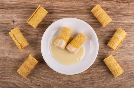 Wafer rolls with condensed milk in saucer, few wafer rolls on wooden table. Top viewの写真素材