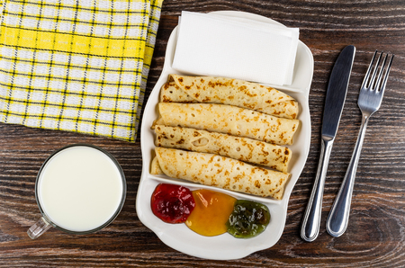 Checkered napkin, cup of milk, partitioned dish with different jams, pancake rolls, tissue, fork and knife on dark wooden table. Top viewの写真素材
