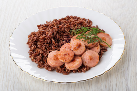 White glass plate with red boiled rice and pieces of fried sausages on wooden tableの写真素材