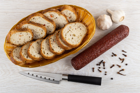 Wicker basket with pieces of bread, smoked sausage, garlic, black pepper, clove spice, kitchen knife on wooden table. Top viewの写真素材