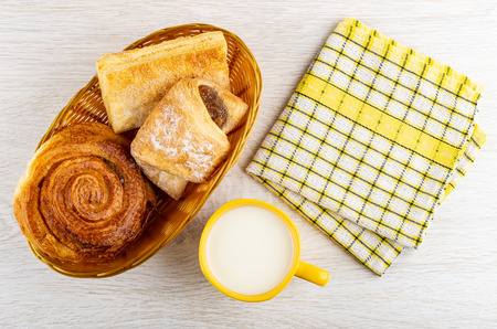 Different sweet pastry in wicker basket, checkered napkin, yellow cup with milk on wooden table. Top...
