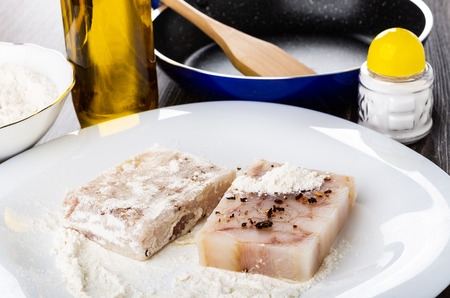 Pressed pieces of pollock fillet in flour with condiment on white dish, bottle of vegetable oil, frying pan, salt shaker on wooden tableの写真素材