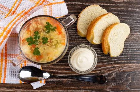 Soup with chicken meat, rice and vegetables in glass bowl, checkered napkin, pieces of bread, bowl with mayonnaise, spoon on wooden table. Top viewの写真素材