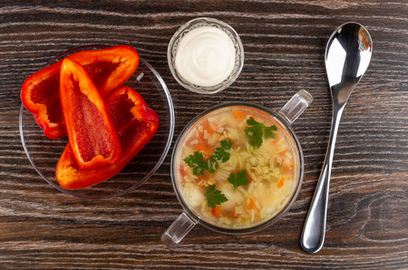 Soup with chicken meat, rice and vegetables in glass bowl, bowl with mayonnaise, pieces of sweet pepper in saucer, spoon on wooden table. Top viewの写真素材
