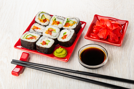 Vegetable rolls with sweet pepper, cucumber, carrot and lettuce, wasabi in red ceramic plate, soy sauce in bowl, pickled ginger in plate, sticks on support on wooden tableの写真素材