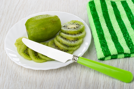Peeled kiwi fruits, slices of kiwi in white glass plate, table knife, striped green napkin on wooden tableの写真素材