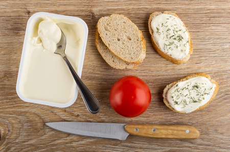 Spoon in white plastic jar with melted creamy cheese, pieces of bread, tomato, sandwiches with cheese and dill, kitchen knife on wooden table. Top viewの写真素材