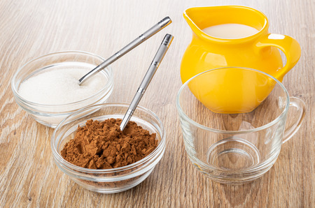 Cacao powder, teaspoon in bowl, teaspoon in bowl with sugar, pitcher with milk, empty transparent cup on wooden tableの写真素材