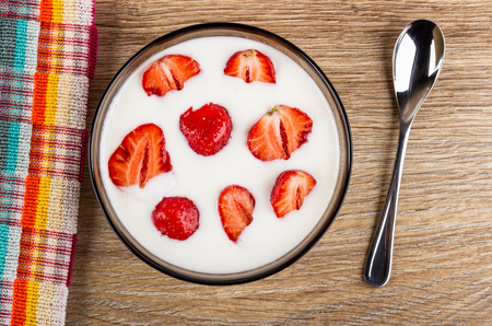 Checkered napkin, pieces of fresh strawberries in brown transparent bowl with yogurt, teaspoon on wooden table. Top viewの写真素材
