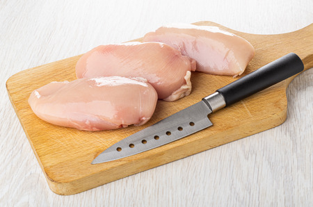 Pieces of raw chicken breast, kitchen knife on cutting board on wooden tableの写真素材