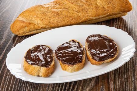 Loaf of bread, sandwiches with chocolate melted cheese in white dish on wooden table