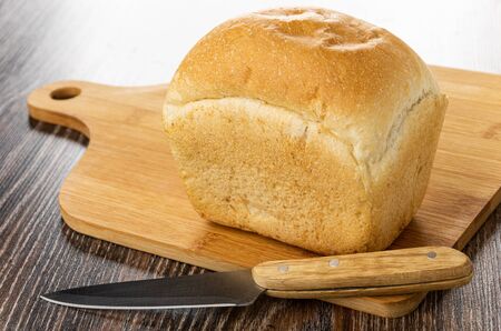 Small loaf of wheaten bread on bamboo cutting board, kitchen knife on dark wooden table