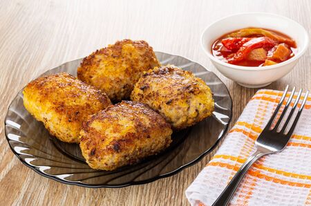 Fried patties in transparent brown plate, bowl with lecho, fork on checkered napkin on wooden table