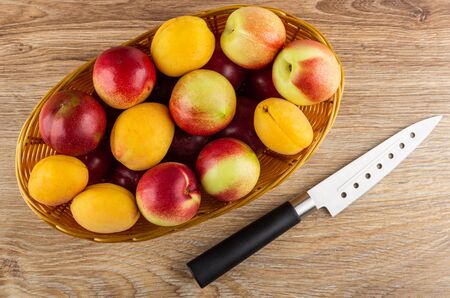 Heap of plums, apricots and nectarines in wicker basket, kitchen knife on wooden table. Top viewの写真素材