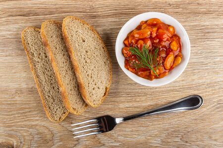 Slices of bread, beans with vegetables and tomato sauce, dill in white bowl, fork on wooden table. Top viewの写真素材