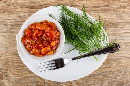 Beans with vegetables and tomato sauce in white bowl, fresh dill, fork on glass dish on wooden table. Top viewの写真素材