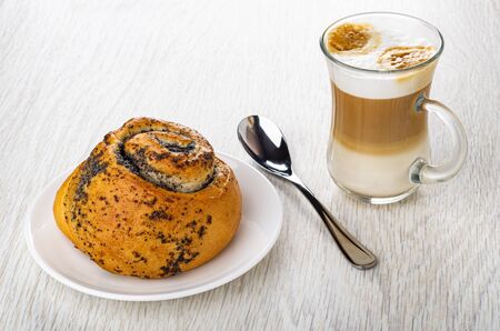 Sweet bun with poppy in white saucer, spoon, latte-macchiato in transparent cup on wooden tableの写真素材