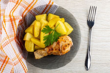 Checkered napkin, grilled chicken leg with fried potato, parsley in gray transparent plate, fork on light wooden table. Top viewの写真素材