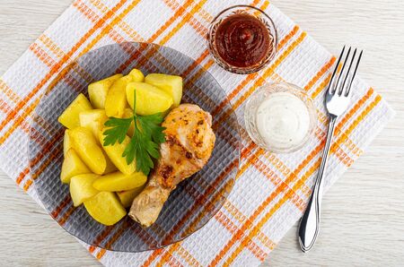 Grilled chicken leg with fried potato, parsley in gray transparent plate, bowls with ketchup and mayonnaise, fork on checkered napkin on light wooden table. Top viewの写真素材